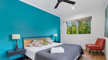 Un dormitorio moderno con una pared verde azulada, una cama con colcha gris, dos lámparas de noche, una silla roja y una ventana con vistas a la vegetación.