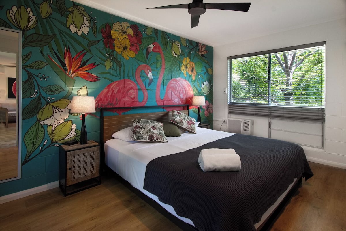A bedroom with a large bed, floral wall art, a ceiling fan, and a window with blinds.
