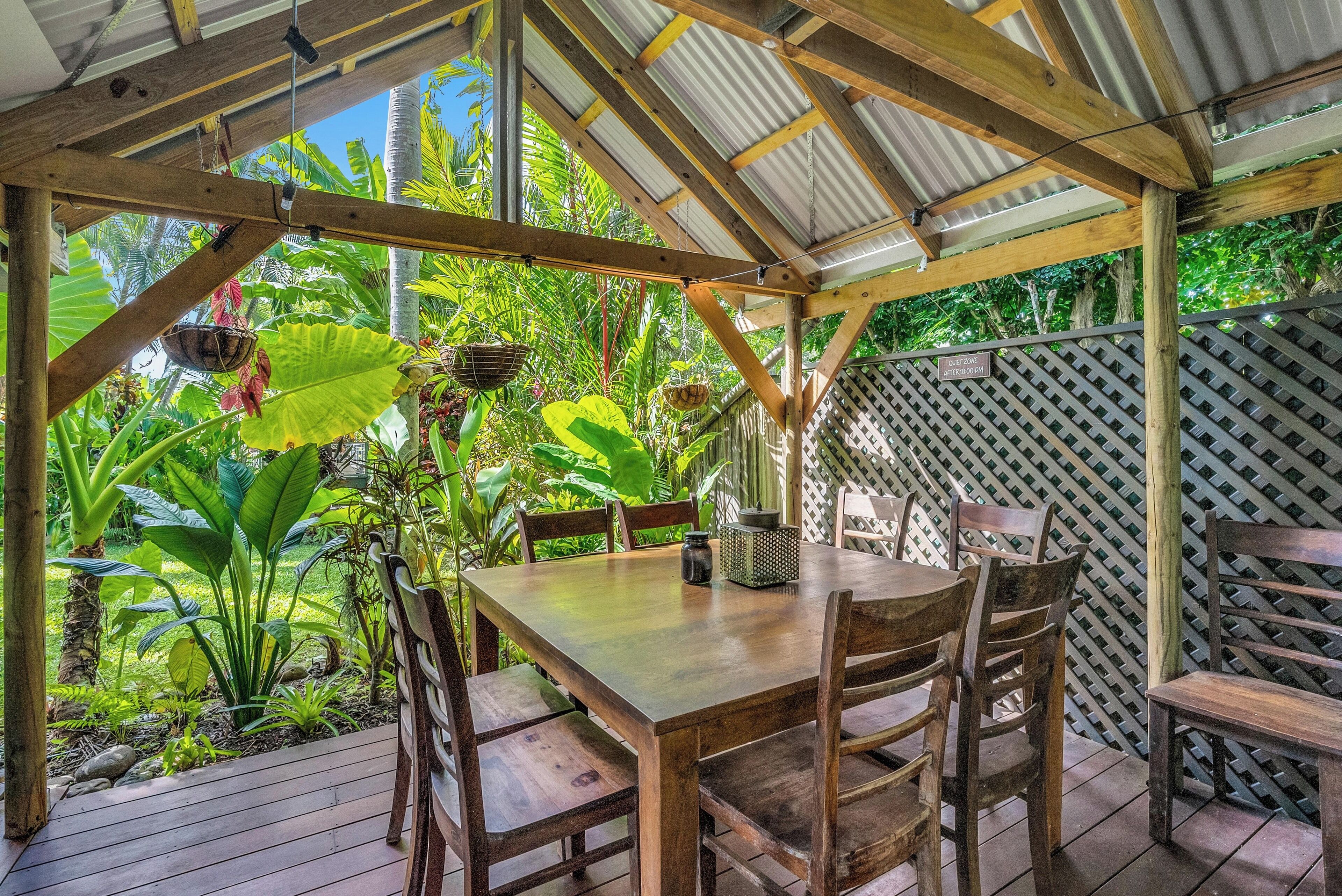 A wooden table and chairs under a metal roof, surrounded by lush tropical plants.
