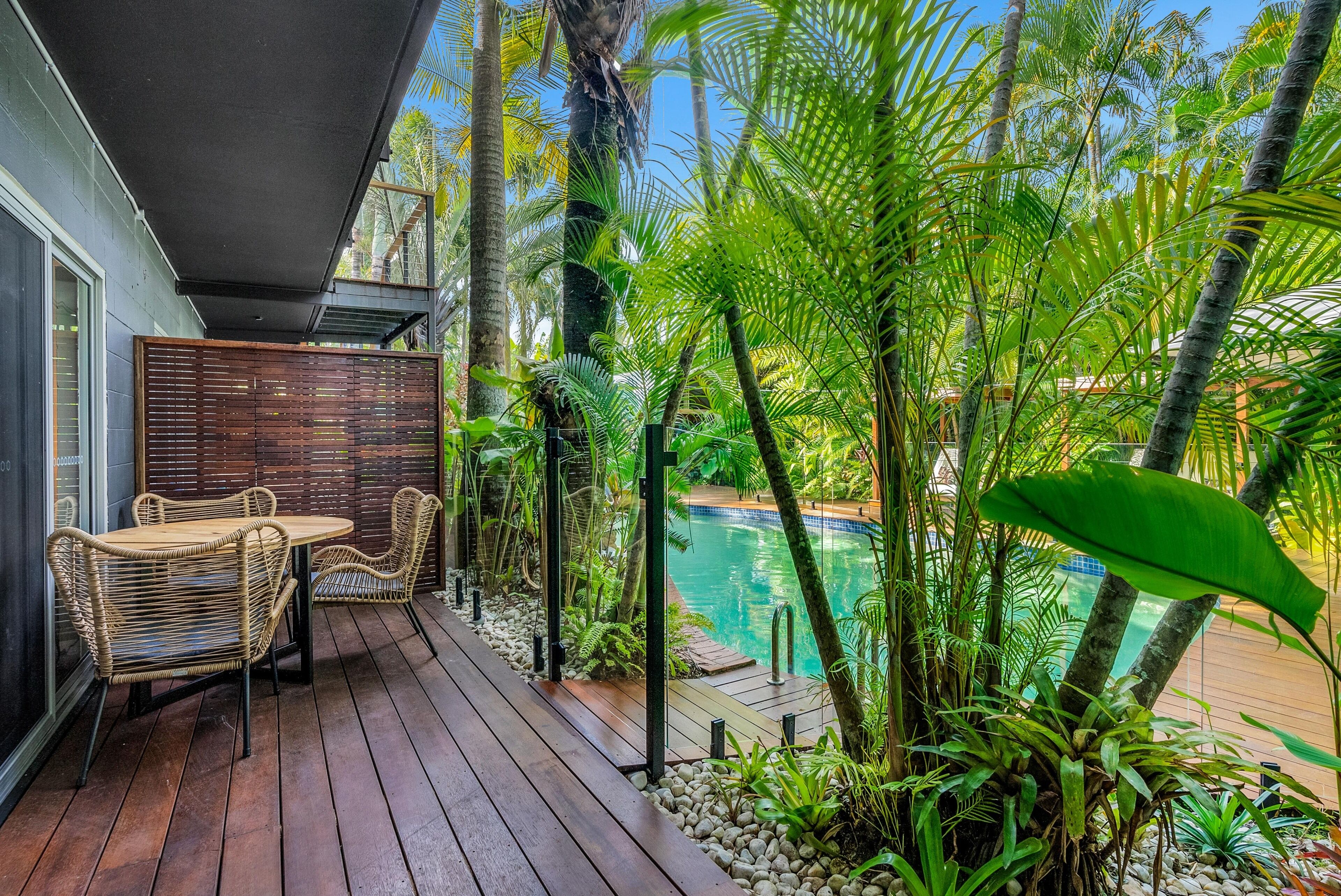 Une terrasse en bois avec des meubles en osier, une piscine et des plantes tropicales.