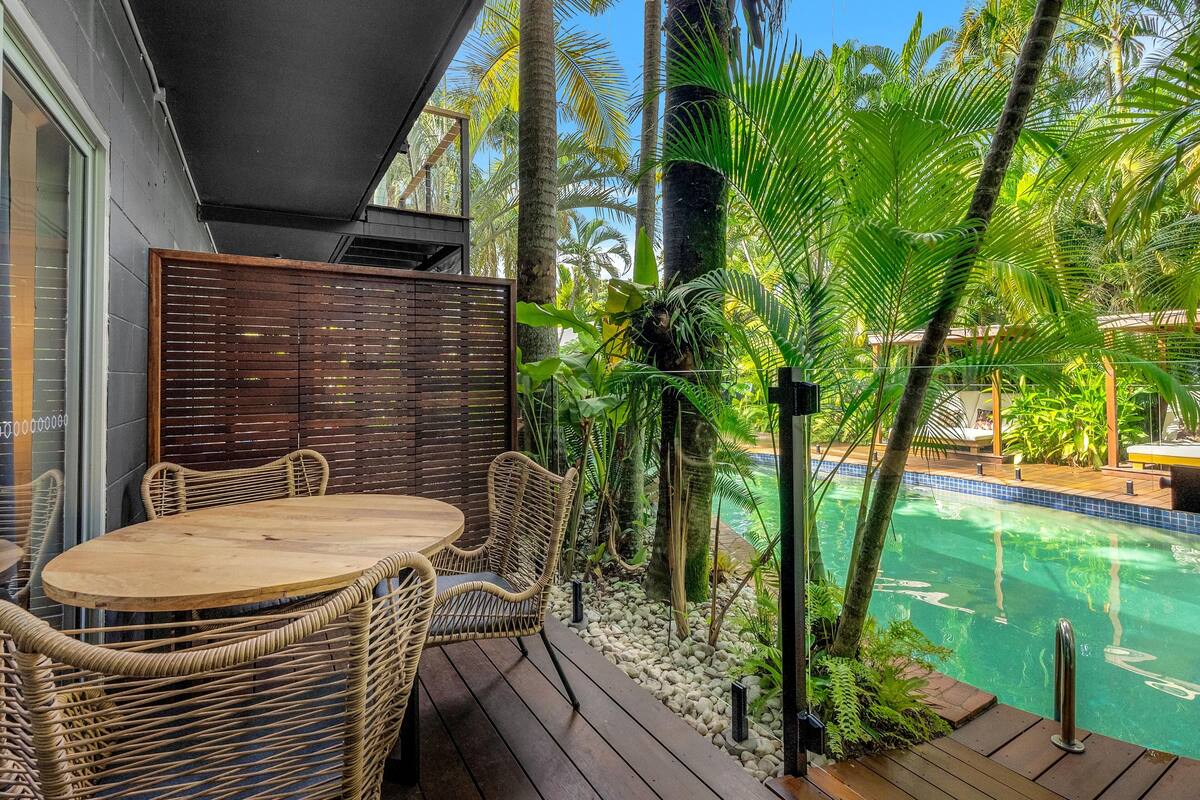A wooden deck with a round table and wicker chairs, overlooking a swimming pool surrounded by tropical plants.