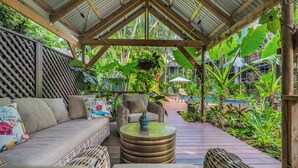 A covered outdoor seating area with wicker furniture, a round table, and a view of a pool and lush greenery. - The Palms at Palm Cove (Palm Cove)
