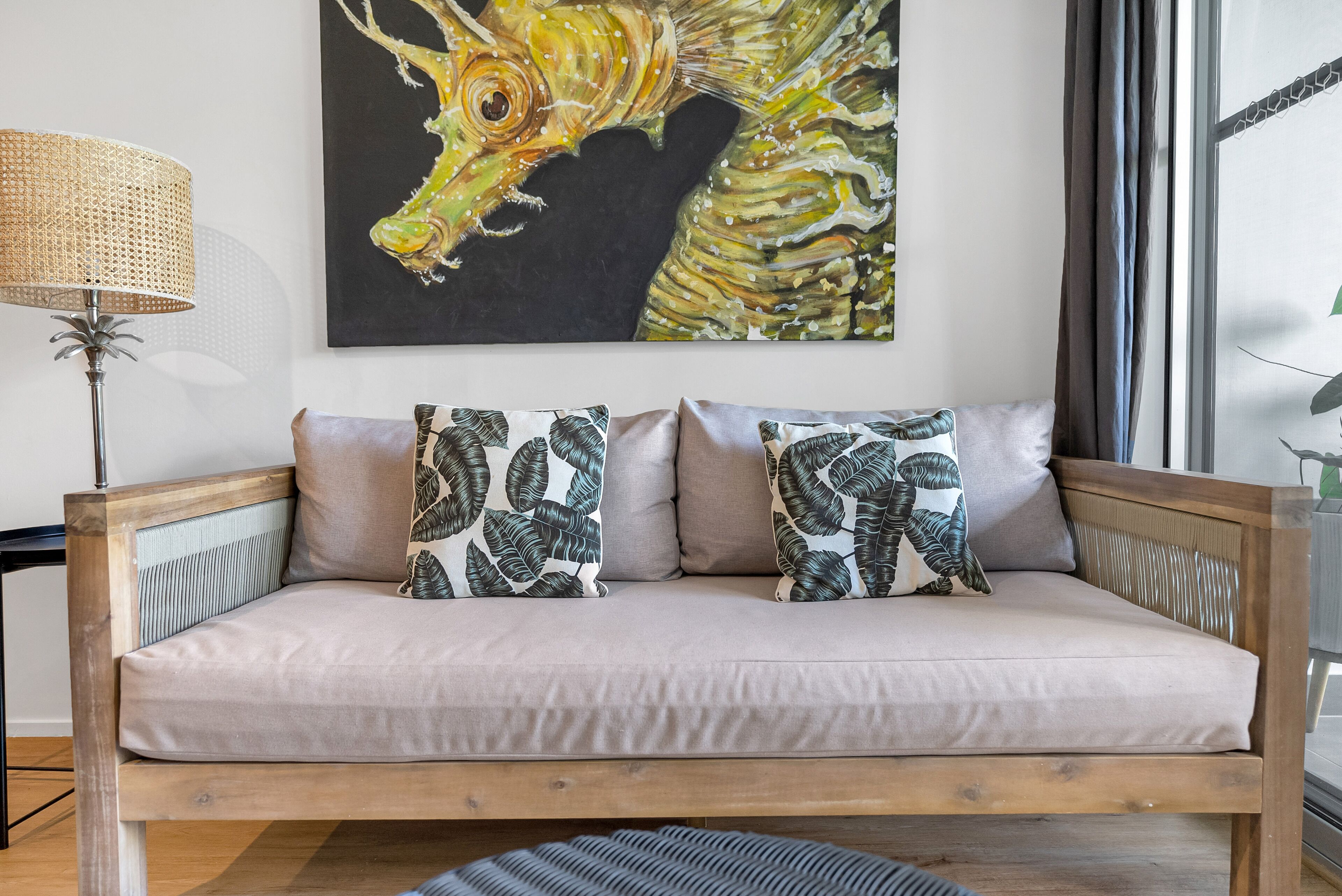 A wooden sofa with a light-colored cushion, two patterned pillows, a woven floor lamp, and a large abstract painting on the wall.