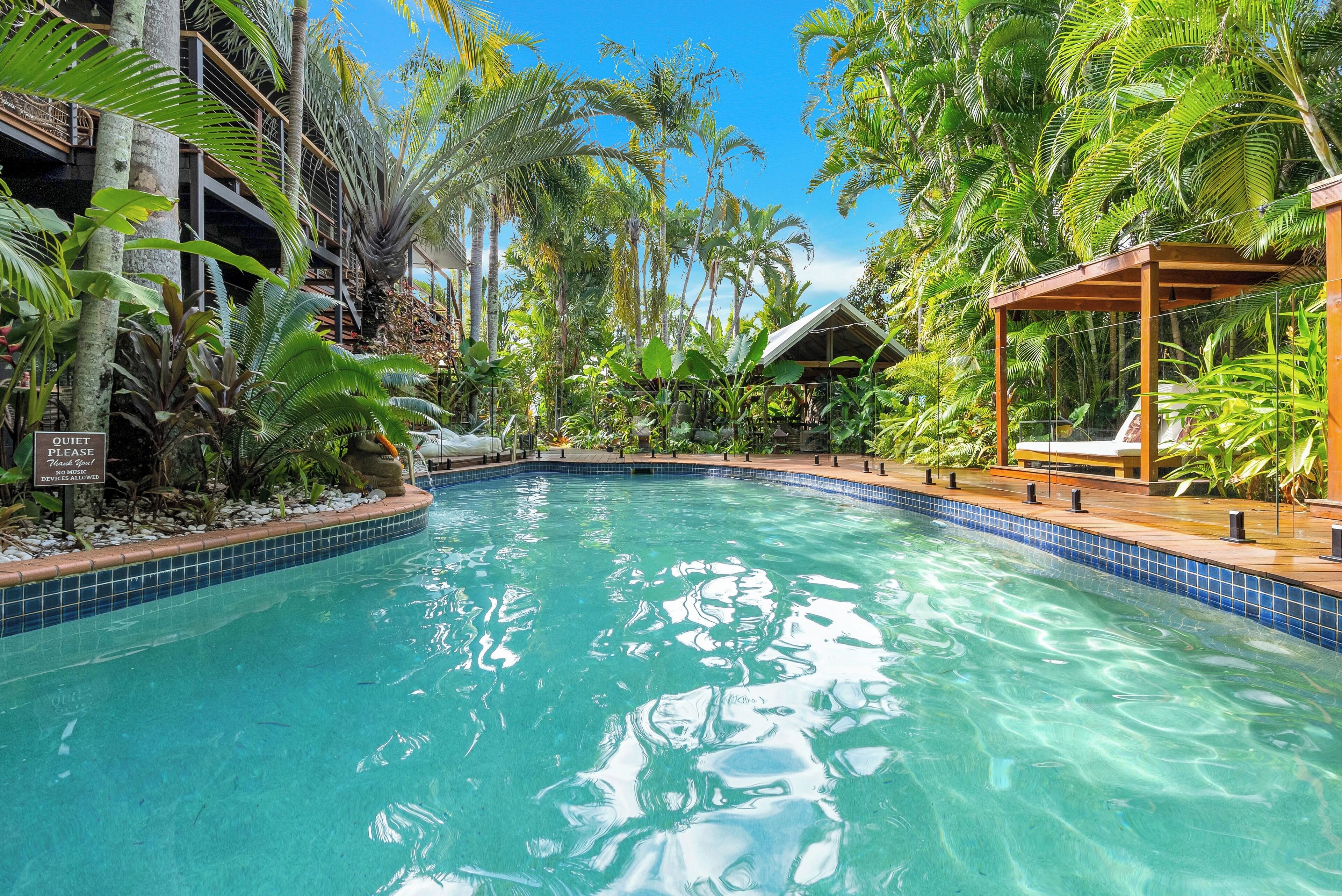 Une piscine aux eaux cristallines, entourée d’une végétation tropicale luxuriante, avec une terrasse en bois et un espace salon abrité.