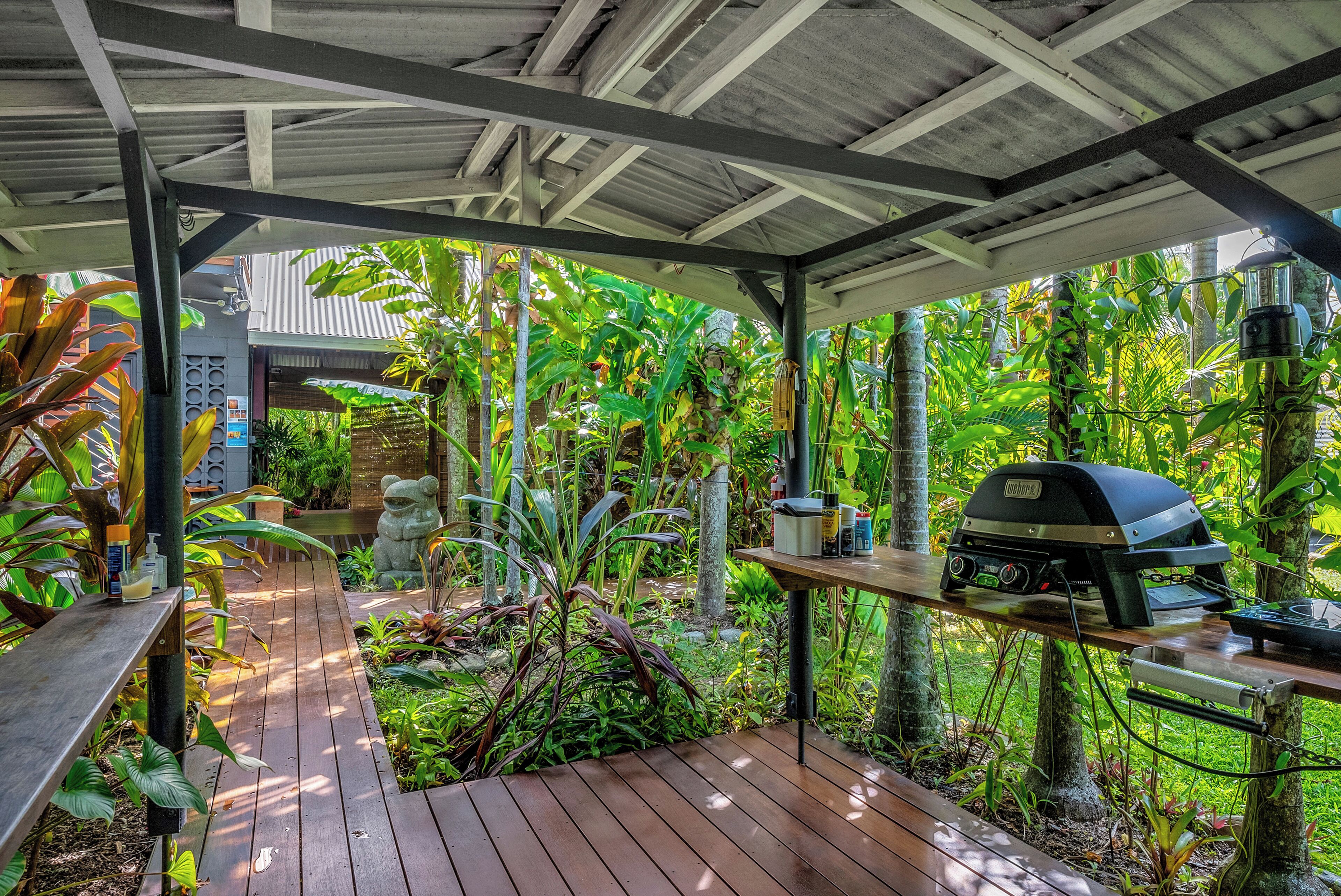A covered outdoor area with a wooden deck, a barbecue grill, and lush tropical plants.