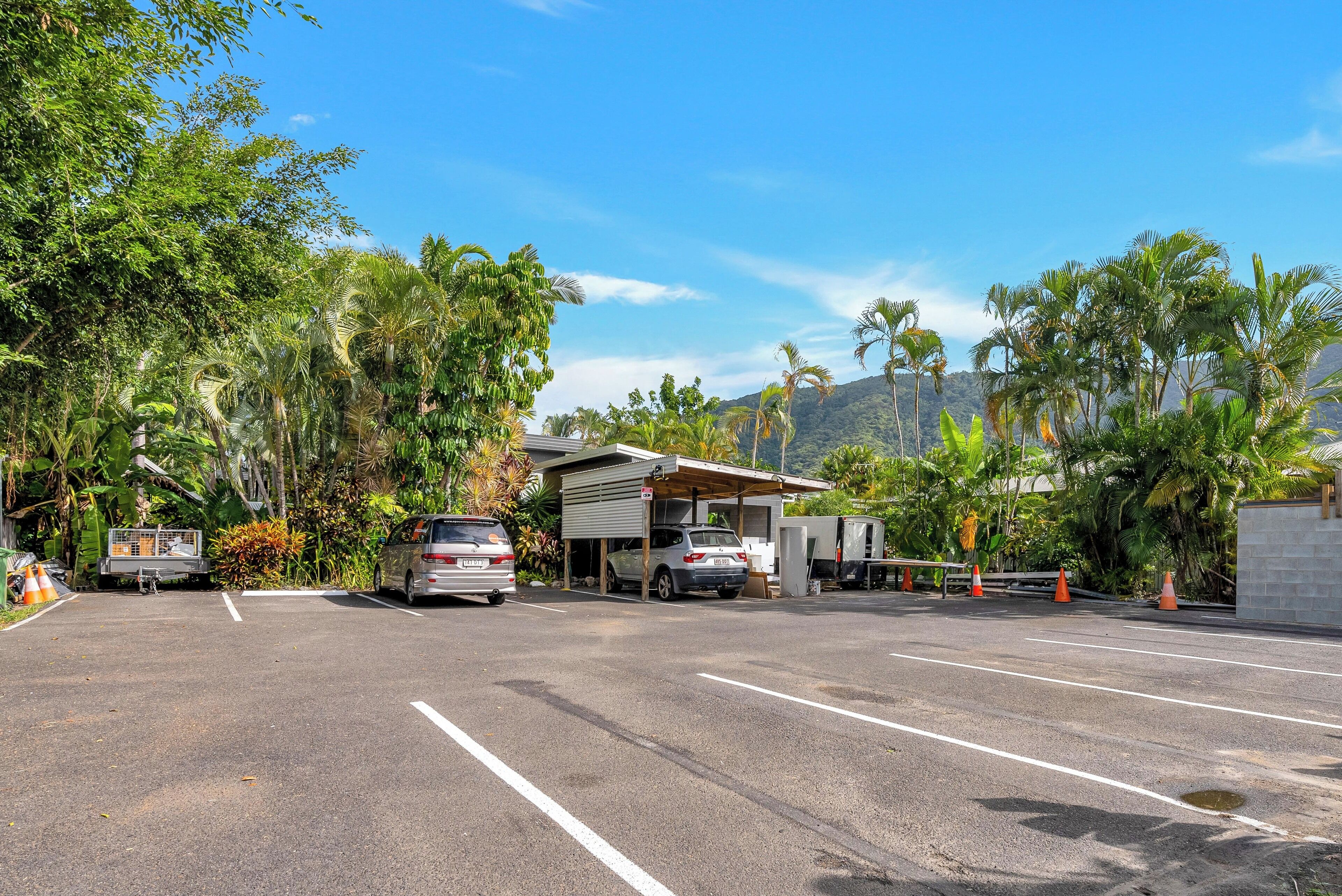 A parking lot with two cars, a building with a covered area, and lush greenery in the background.