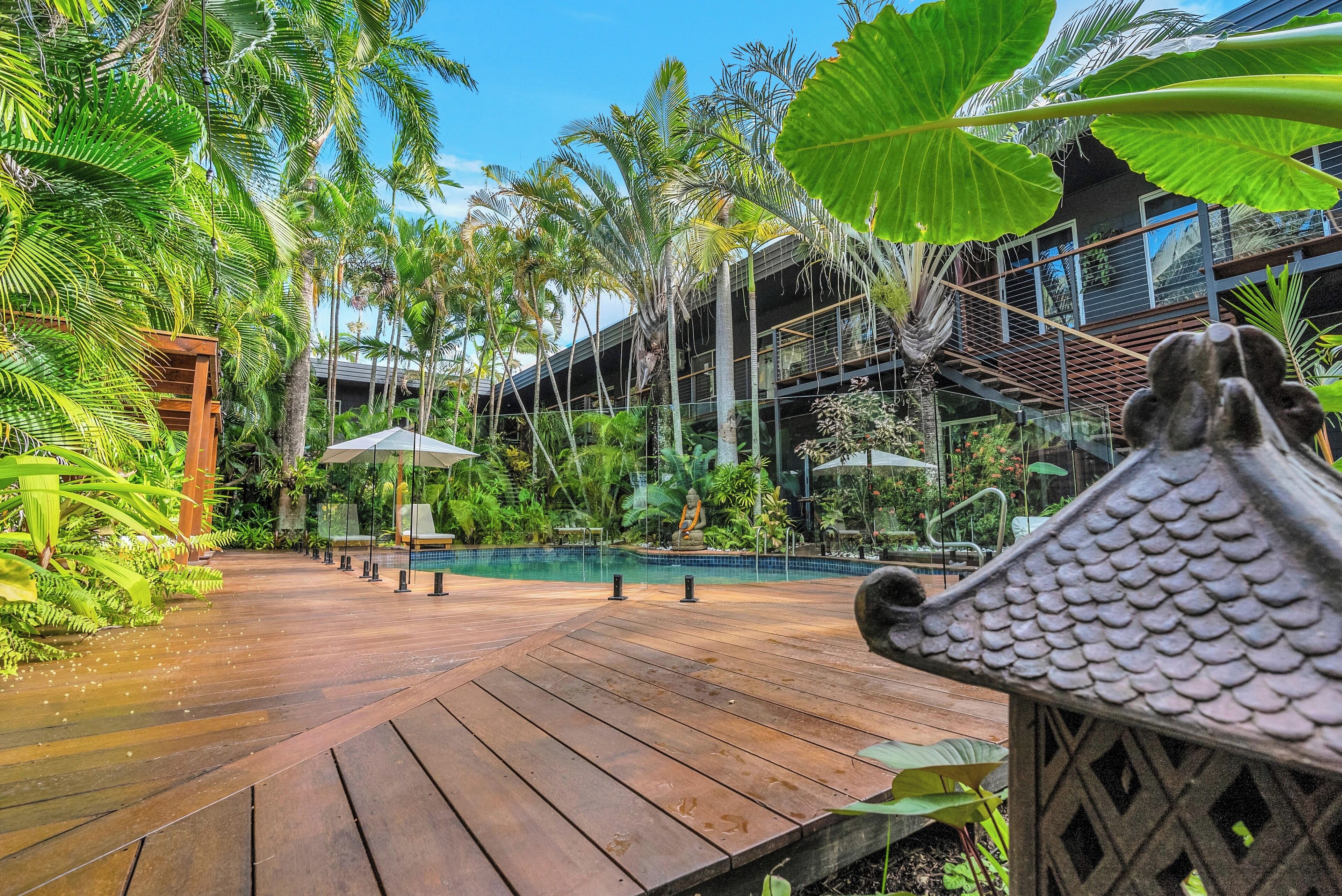 A wooden deck with a pool, surrounded by lush tropical plants and a modern building with balconies.