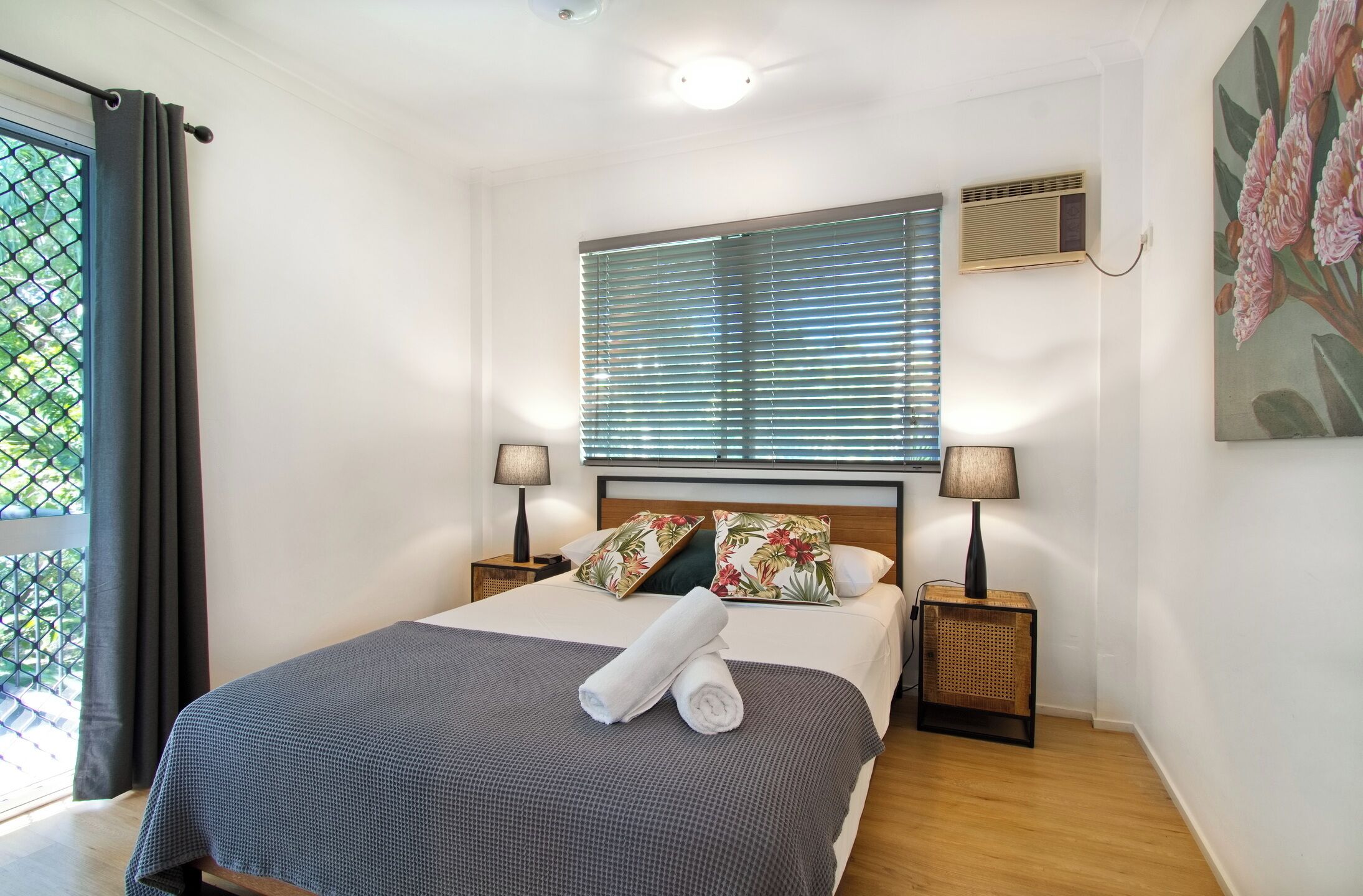 Ein ordentlich bezogenes Bett mit karierter Tagesdecke und zwei Kissen, zwei Nachttischlampen, ein Fenster mit Jalousien, eine Wand-Klimaanlage und ein Blumenbild.