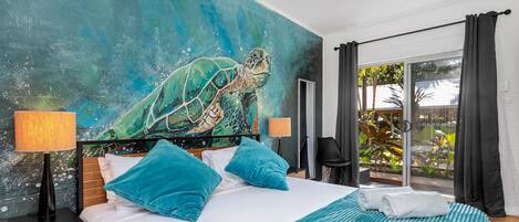 Un dormitorio con un gran mural de una tortuga marina, una cama con ropa de cama azul, dos lámparas de noche y vistas a un jardín a través de una puerta corredera.