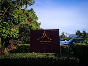 Señal de Khum Nakhon con un diseño moderno, rodeada de exuberante vegetación y automóviles estacionados.