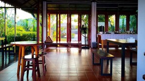 A spacious room with a tiled floor, a wooden table, and a bench. There are large windows with a view of greenery outside. - Khum Nakhon Hotel (Nakhon Si Thammarat)