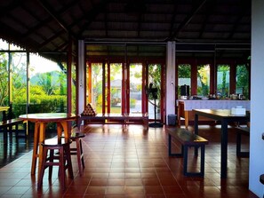 A spacious room with a tiled floor, a wooden table, and a bench. There are large windows with a view of greenery outside. - Khum Nakhon Hotel (Nakhon Si Thammarat)
