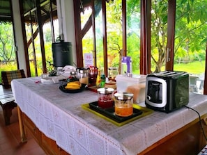 A breakfast table with a toaster, jars of jam, a fruit tray, and a coffee maker. - Khum Nakhon Hotel (Nakhon Si Thammarat)