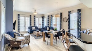 Un salon spacieux comprenant un coin repas, deux canapés, un ventilateur de plafond et de grandes fenêtres avec des rideaux.