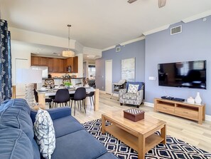 Una sala de estar moderna con sofá, mesa de centro y televisión. Se aprecia un comedor con sillas y mesa, y una cocina al fondo.