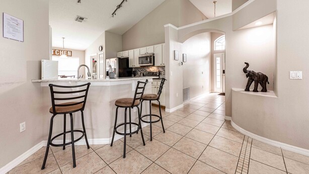 A kitchen with white cabinets, a microwave, and a refrigerator. There are two bar stools with cushioned seats. The floor is tiled with beige tiles. - Premier Host-Beautiful 4BR/2BA Pool HEAT Home SAFE QUIET Lot - Disney 15 Minutes (Kissimmee)