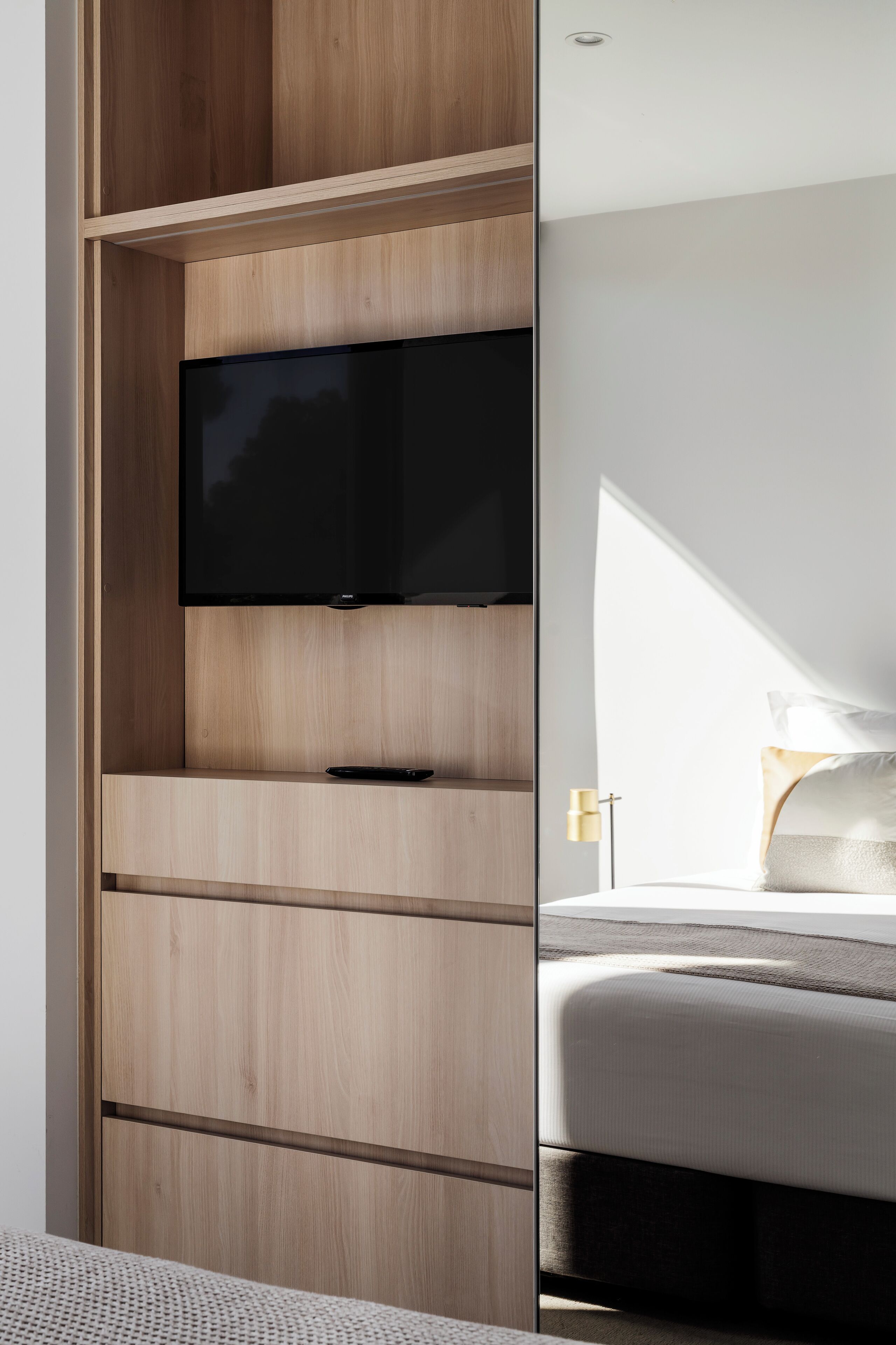 Habitación moderna con televisión integrada, almacenaje de madera, cama con ropa blanca y ventana que proporciona luz natural.