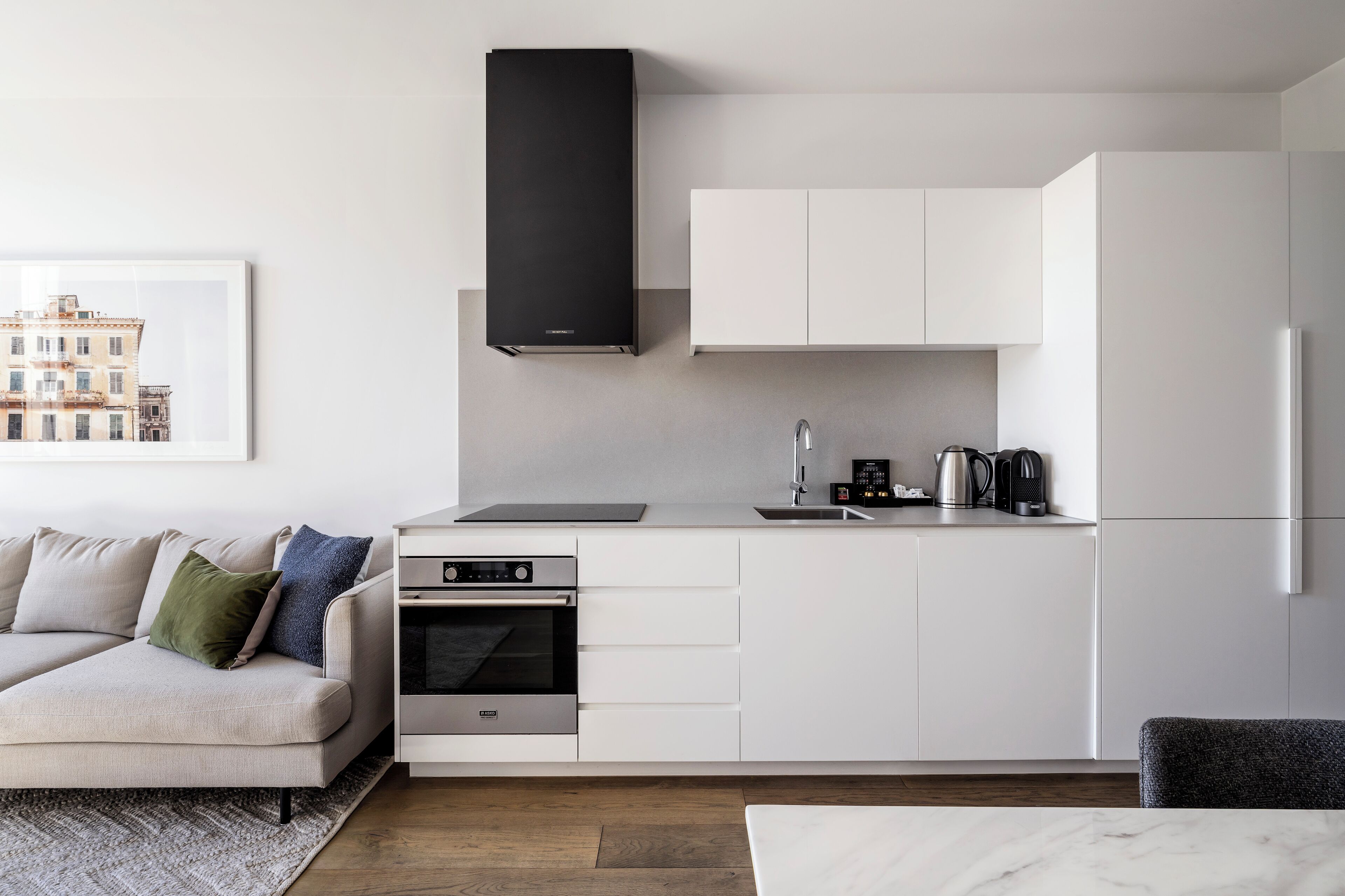 A modern kitchen with white cabinetry, a built-in oven, a black range hood, and a coffee maker on the countertop.