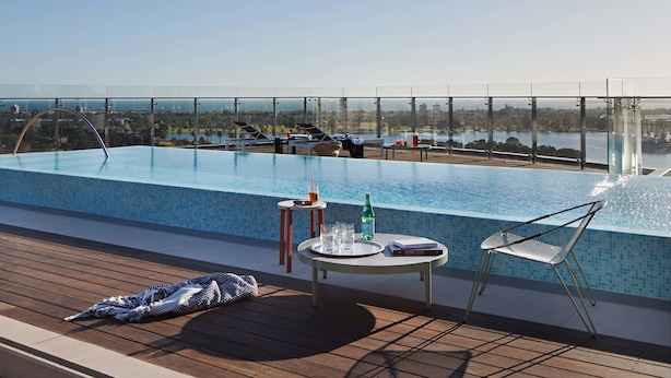 Une piscine sur le toit offrant une vue dégagée sur les gratte-ciel de la ville, une chaise longue, une petite table avec une boisson et une serviette sur la terrasse.