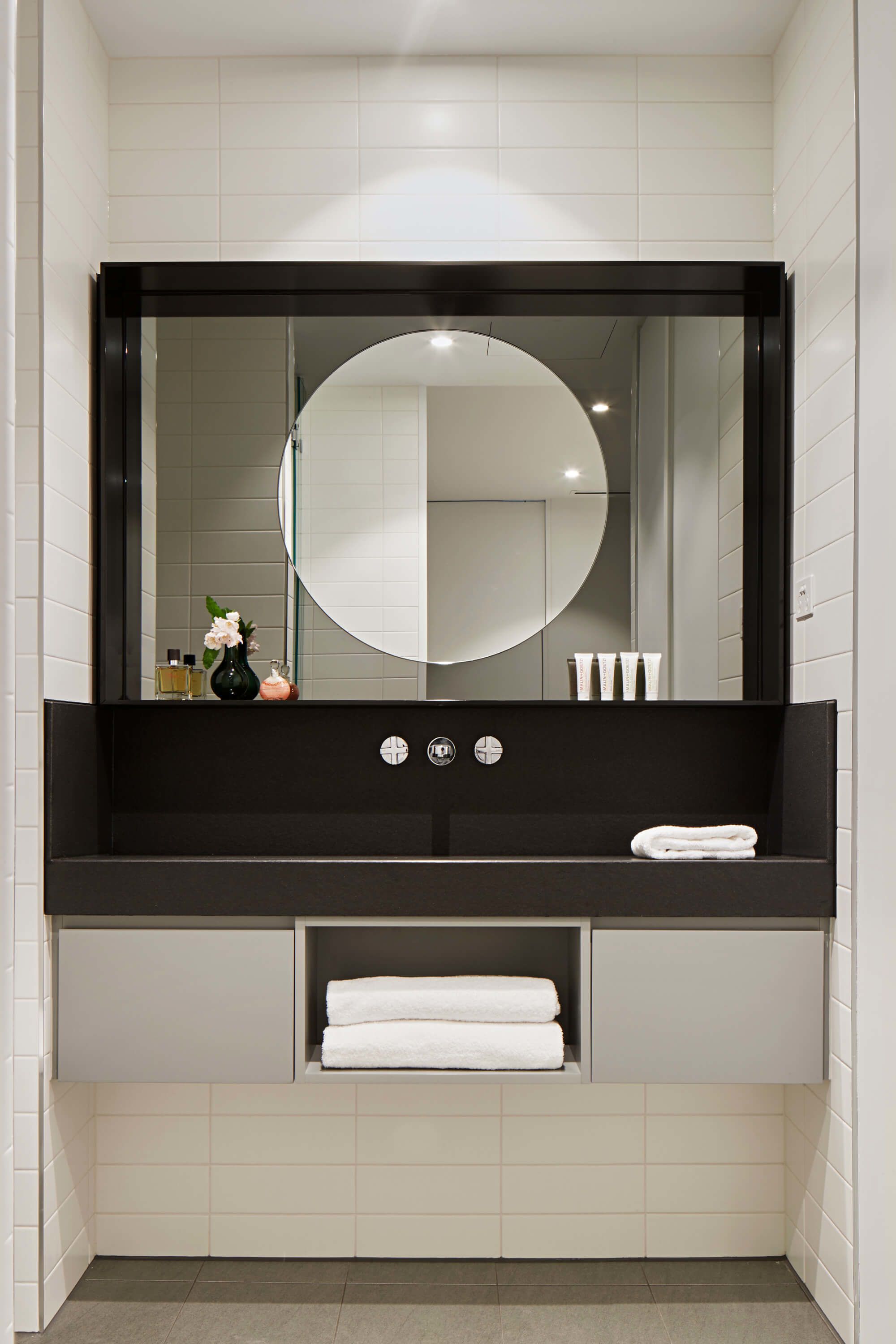 A modern bathroom with a black vanity, a round mirror, and a built-in shelf with toiletries.
