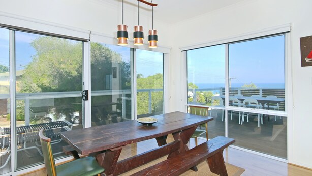 A modern dining area with a wooden picnic table, green chairs, and a large window offering a view of the sea. - Kiltevna (Maslin Beach)