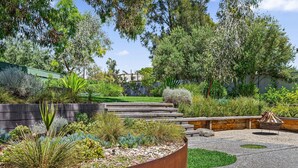 A landscaped garden with a variety of plants, a fire pit, and a wooden staircase leading to a house. - Port of Call (Port Willunga)