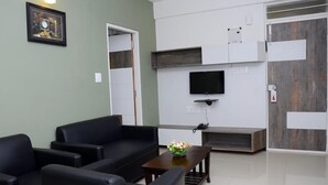Una sala de estar moderna con un sofá de cuero negro, una mesa de centro de madera con un florero, un televisor de pantalla plana montado en una pared blanca y un mueble de madera empotrado.