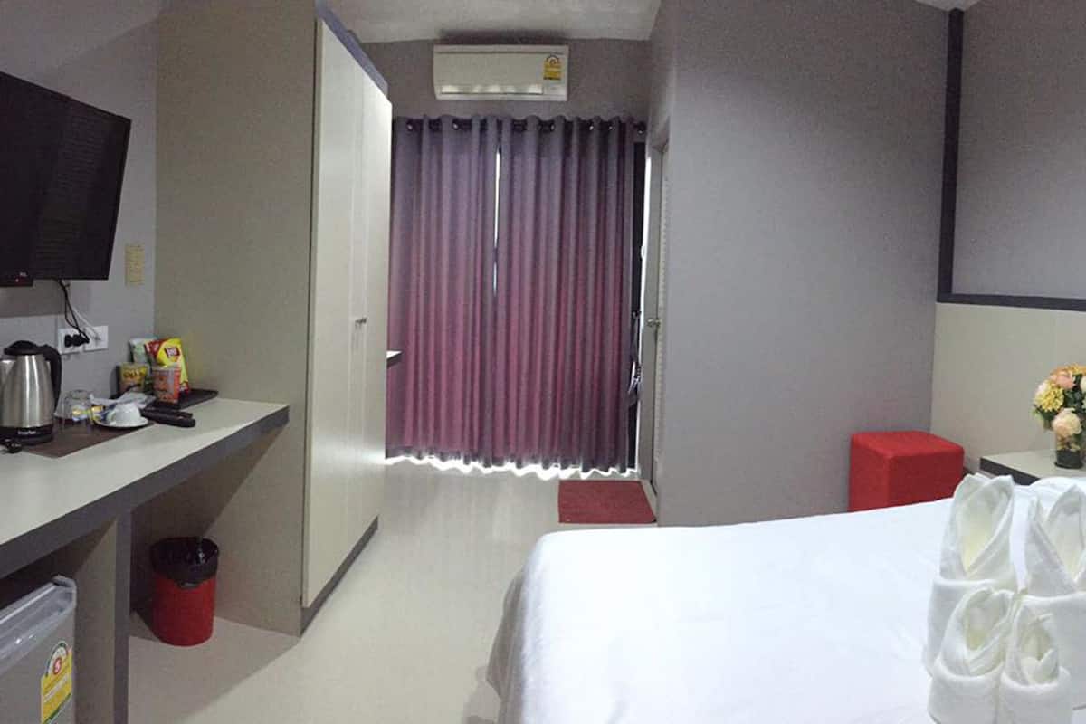 Une chambre d’hôtel avec un lit, une télévision, un bureau avec une bouilloire, une poubelle et un tabouret rouge.