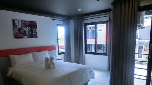 Une chambre d’hôtel moderne avec un grand lit, une tête de lit rouge et une vue sur la ville par la fenêtre.