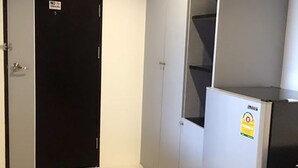 Une pièce avec une porte noire, des murs blancs et un sol blanc. On y trouve un meuble blanc avec une étagère et un réfrigérateur orné d’un autocollant.
