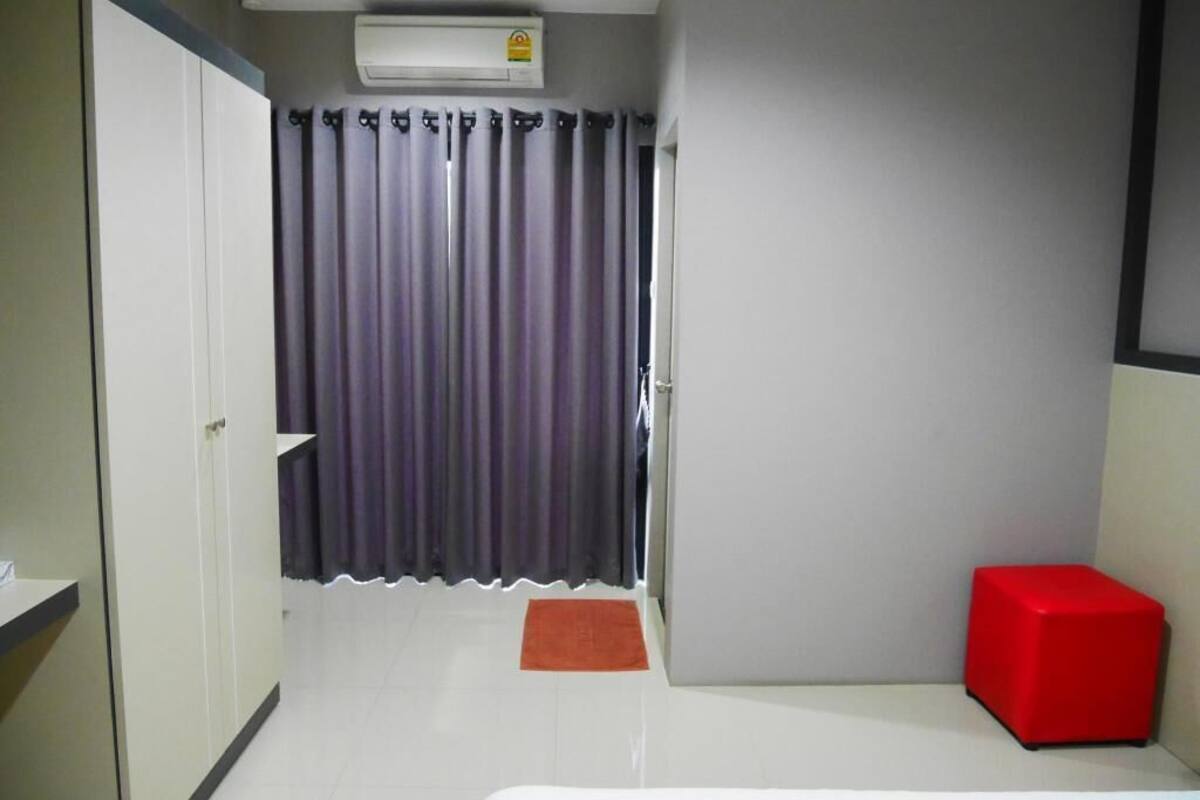 Une chambre d’hôtel avec un lit blanc, un tabouret rouge, un rideau gris et un appareil de climatisation blanc.