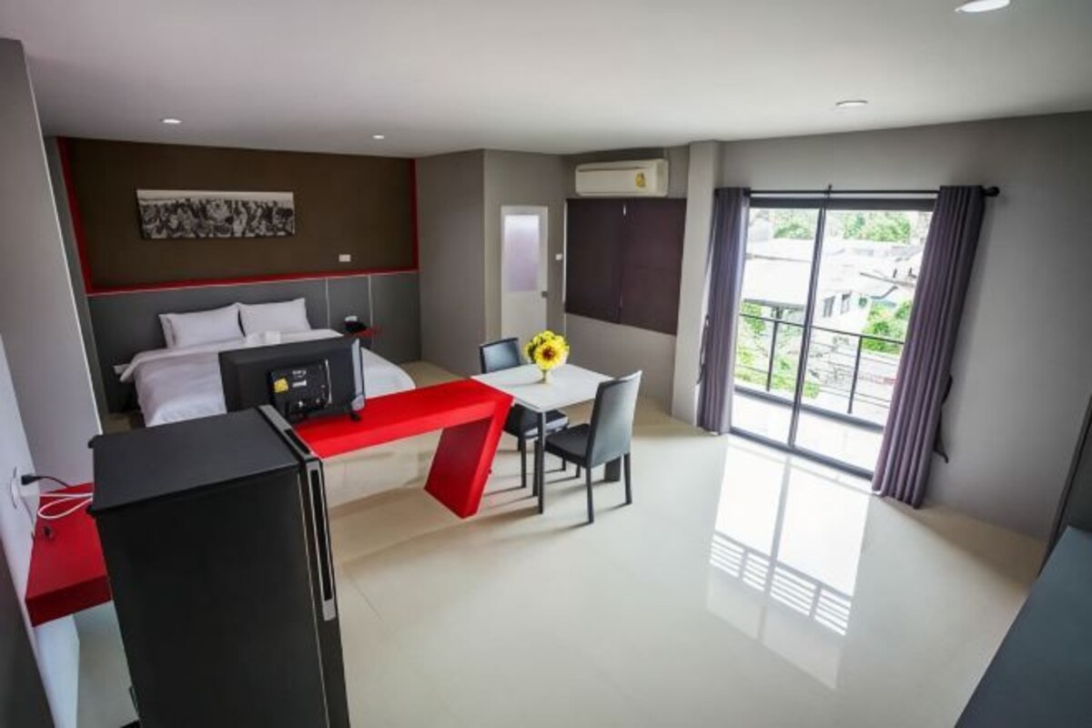 Une chambre d’hôtel comprenant un lit, un bureau avec une chaise, un réfrigérateur, une fenêtre avec des rideaux et une vue sur la verdure environnante.