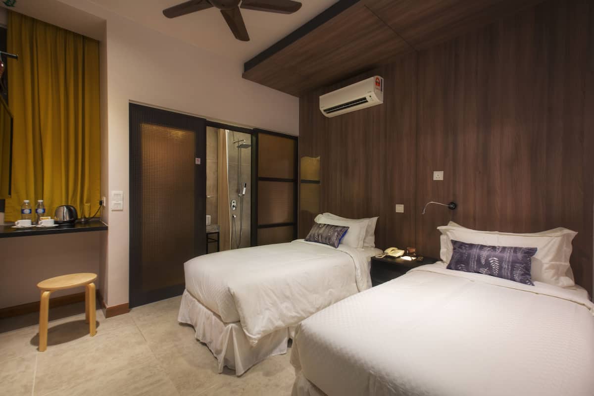 A hotel room with two beds, a small wooden stool, a desk with a chair, and a shower area.