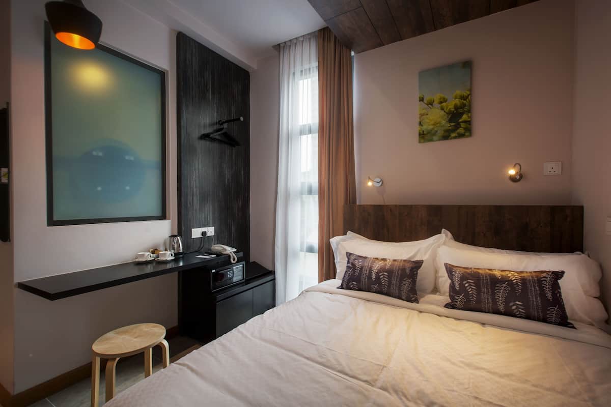 A hotel room with a bed, a desk, a chair, a nightstand, a painting, and a window with curtains.