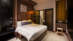 Quarto de hotel moderno com uma cama grande, cabeceira de madeira, ventilador de teto, área de chuveiro e um banquinho.