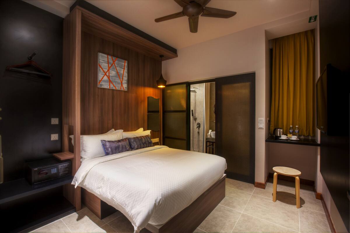 A modern hotel room with a large bed, a wooden headboard, a ceiling fan, a shower area, and a small stool.