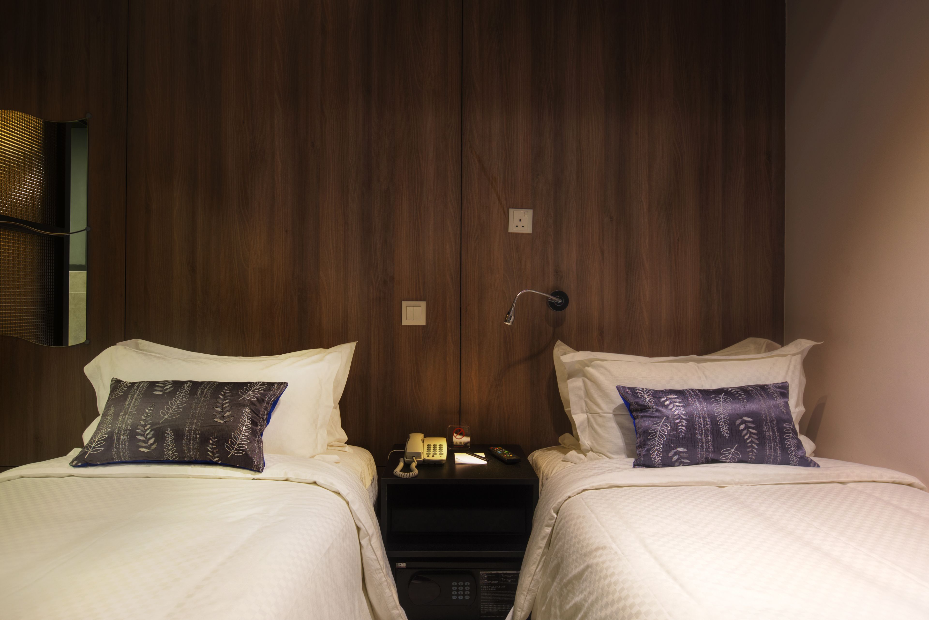 Ett hotellrum med två sängar, var och en med ett vitt överkast och en mörk, mönstrad kudde. Mellan sängarna står ett nattduksbord med en telefon, en fjärrkontroll och en liten lampa.