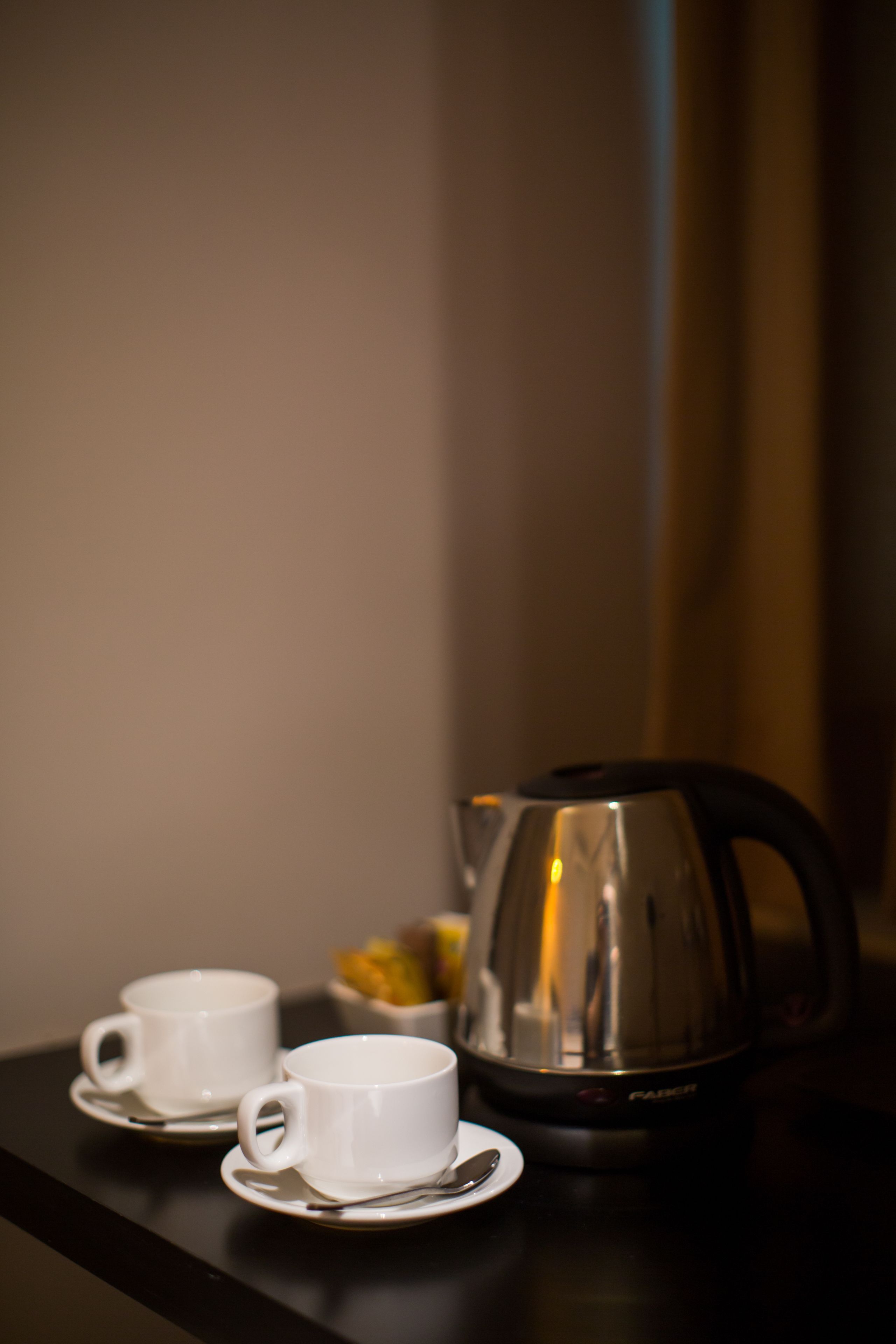 Two white cups on saucers and a silver electric kettle on a dark surface.