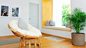 Una habitación con una silla de mimbre, un ventanal con vistas a zonas verdes, una planta en maceta y dos cuadros en la pared.