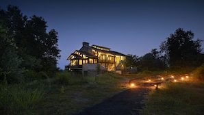 A large, two-story house with a prominent balcony, surrounded by lush greenery and a gravel path with lights. - Pugdundee Safari- Denwa Backwater Escape (Sohagpur)