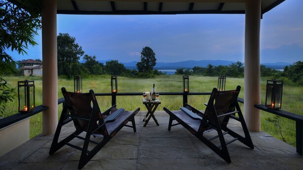 A covered outdoor seating area with two wooden chairs and a table, overlooking a grassy field and distant mountains. - Pugdundee Safari- Denwa Backwater Escape (Sohagpur)