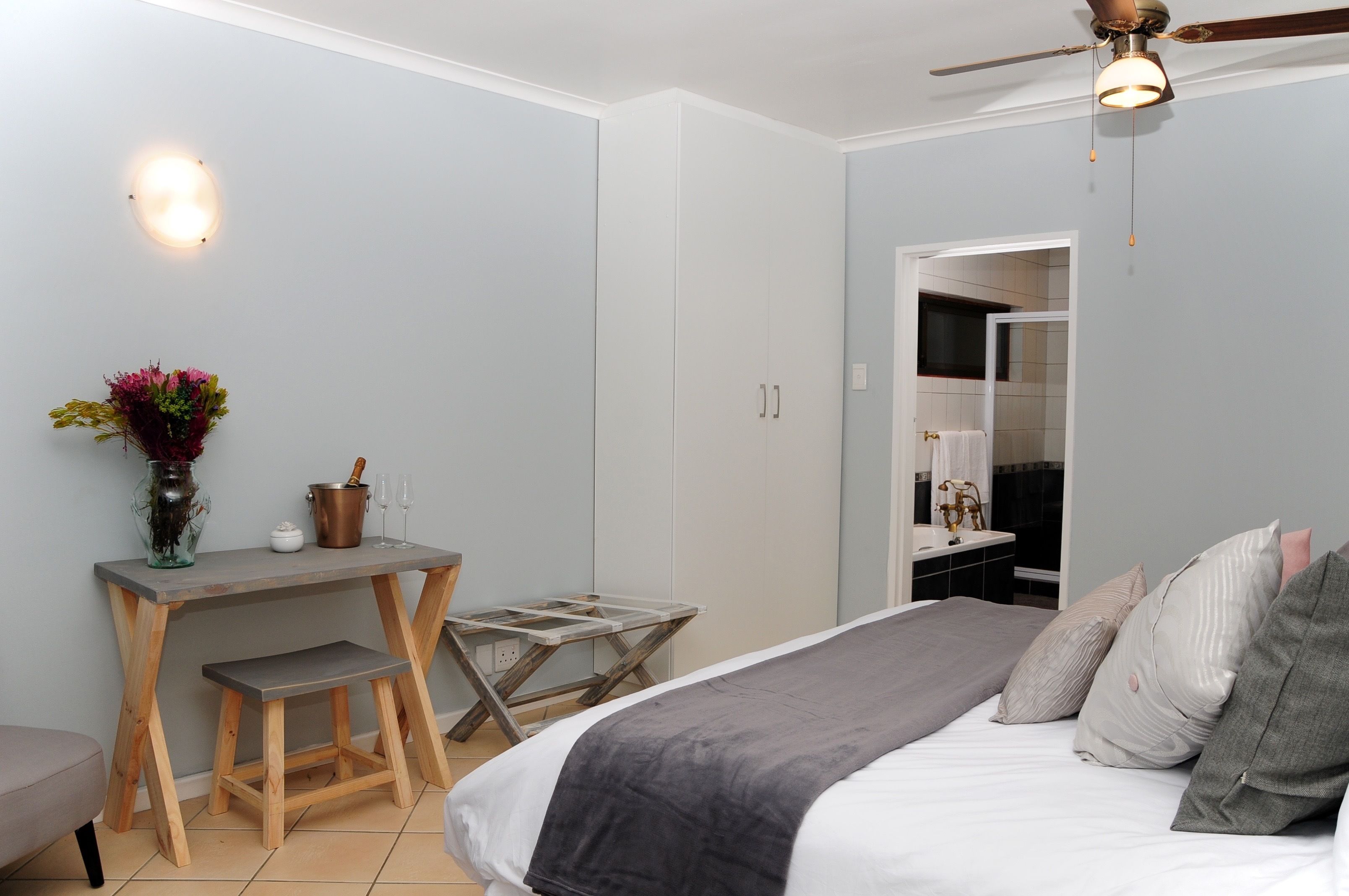 A bedroom with a bed, bedside table, and a desk with a chair. There is a ceiling fan and a wall-mounted light fixture.