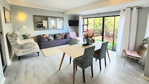 A modern living room with a sofa, dining table, and chairs. There is a TV mounted on the wall and a large window with curtains. - The Salt House (Cape Town)