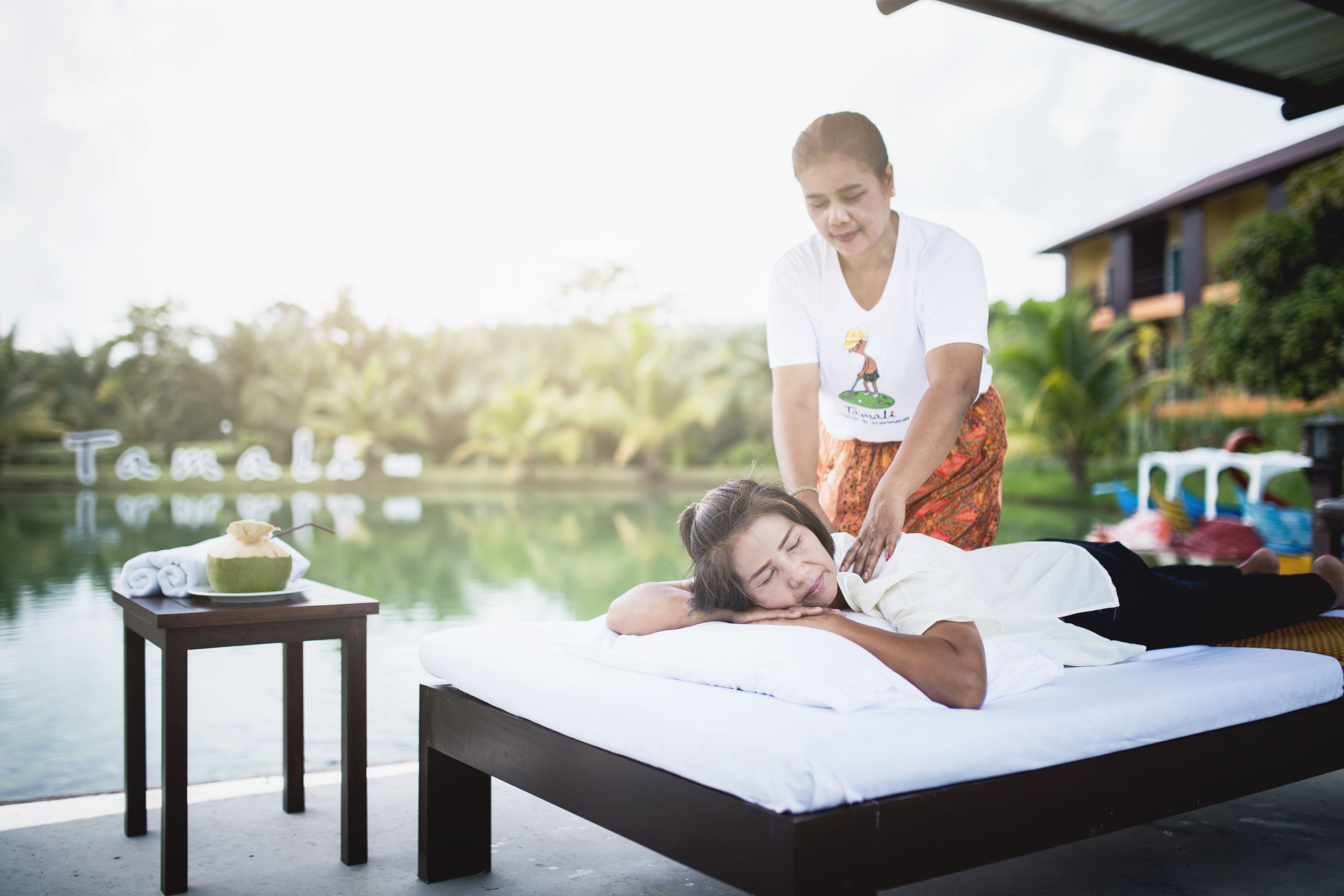 A person lying on a massage table outdoors with a body of water and greenery in the background.