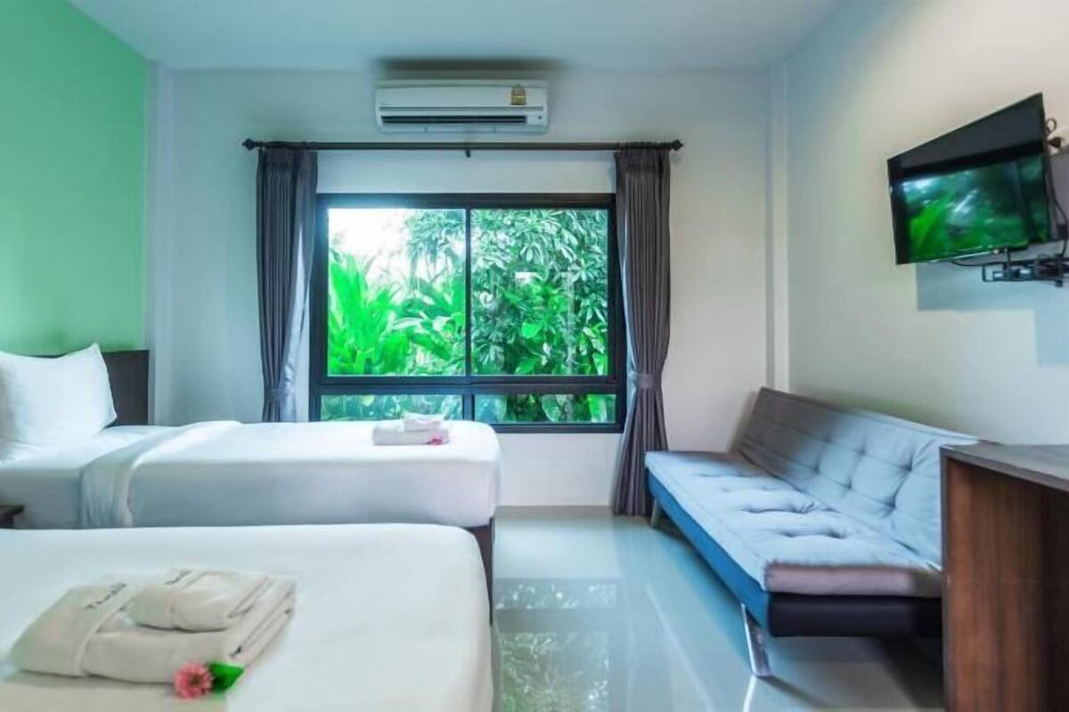 A hotel room with two beds, a sofa, a TV, and a window with a view of greenery.