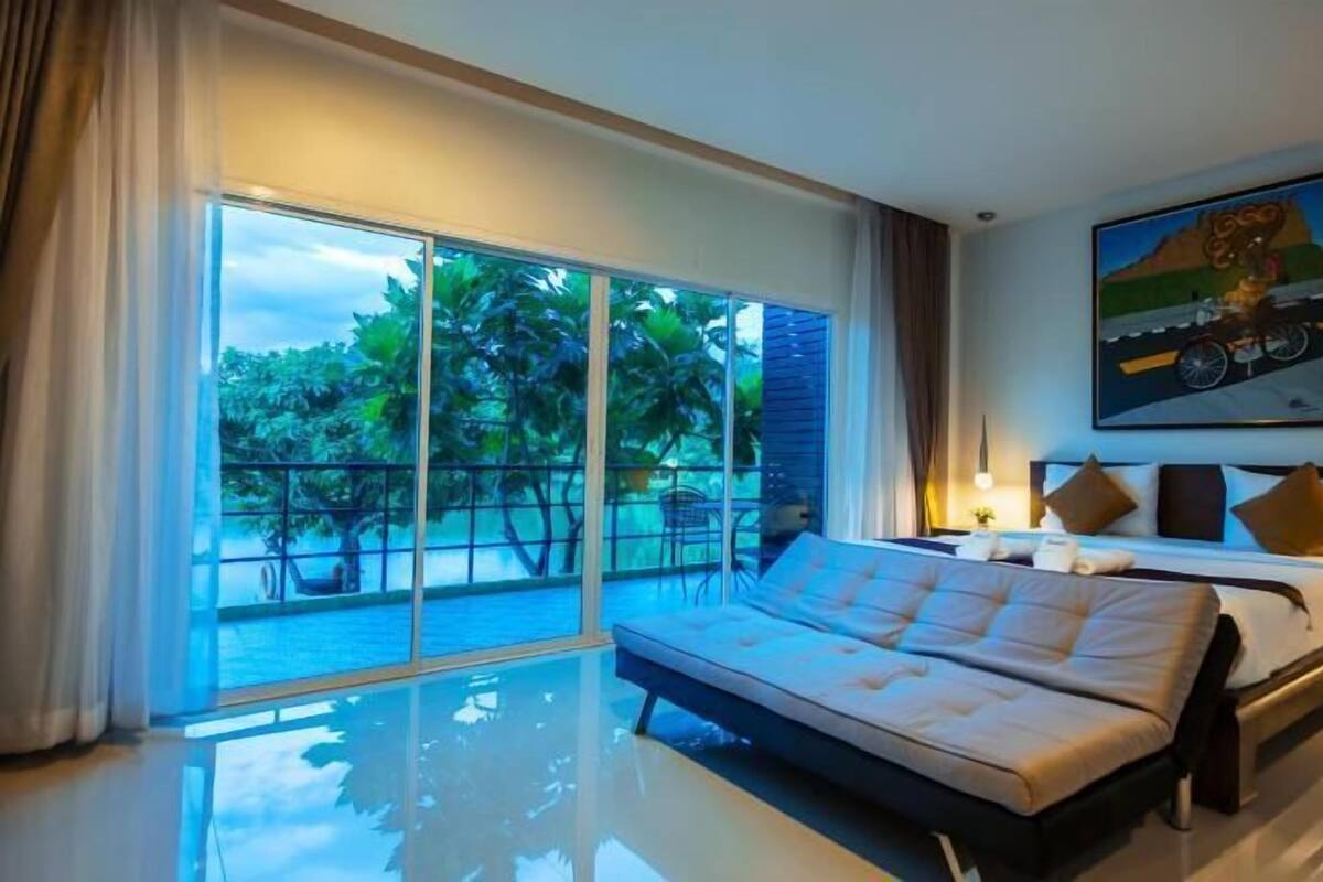 A modern hotel room with a large bed, a balcony with a view of greenery, and a painting on the wall.
