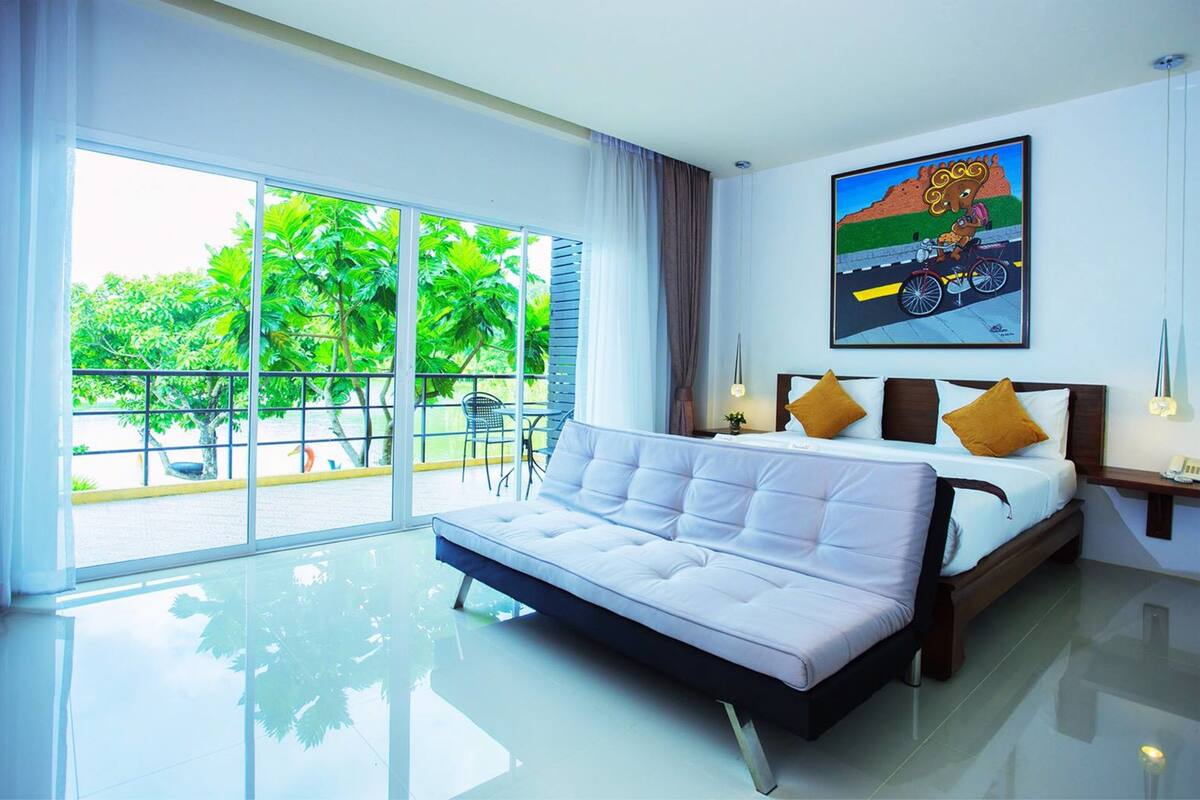 A modern hotel room with a large bed, a balcony with a view of greenery, and a painting on the wall.