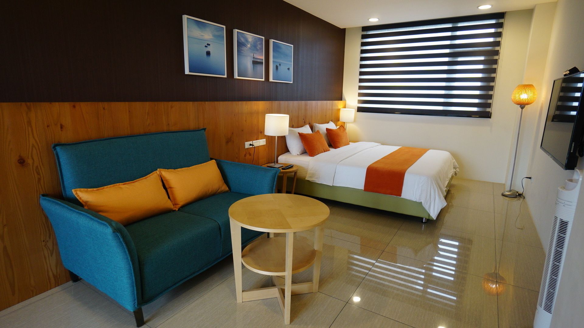Een hotelkamer met een blauwe bank, een bed met een oranje en witte spreideken, een klein rond tafeltje en een aan de muur bevestigde televisie.