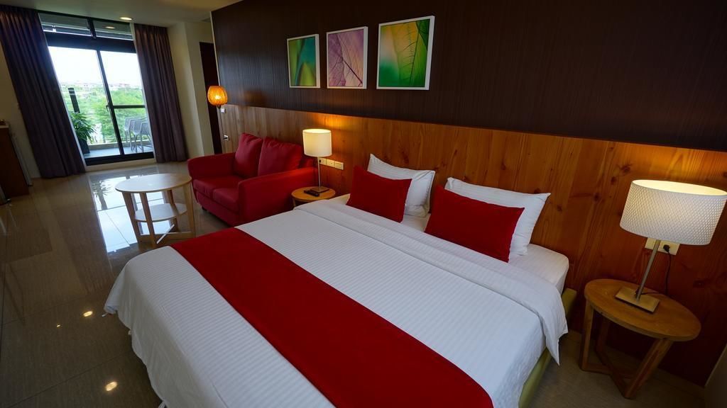 Ett hotellrum med en stor säng, rött och vitt sänglinne, en röd fåtölj, ett litet runt bord och utsikt över grönområden utanför.
