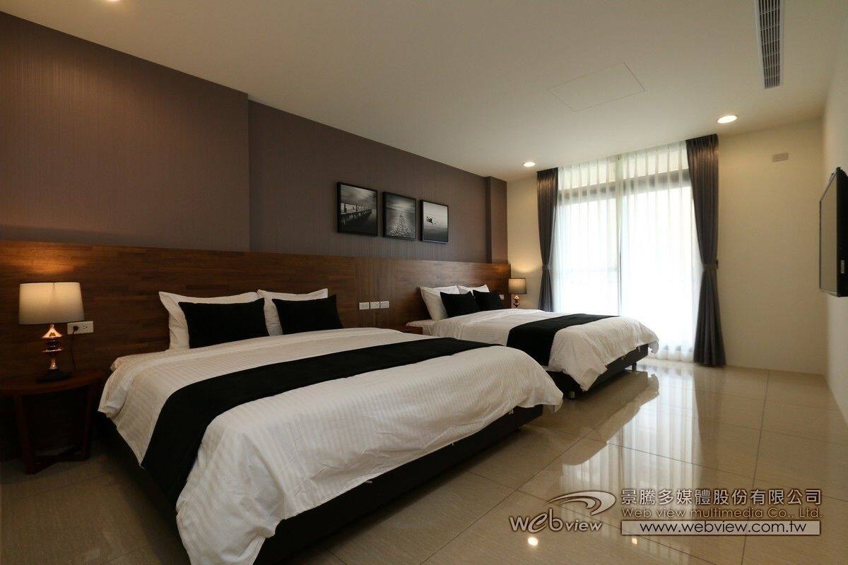 A hotel room with two beds, a wooden headboard, a large window with curtains, and a wall-mounted TV.